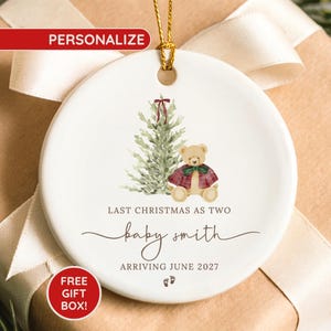 May include: A round white ceramic ornament featuring a watercolor illustration of a green Christmas tree and a teddy bear wearing a red plaid cape. The text reads: LAST CHRISTMAS AS TWO, baby smith, ARRIVING JUNE 2027, with two small baby footprints.