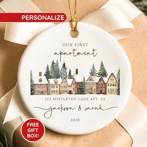 May include: A round white ceramic ornament featuring a watercolor illustration of a row of houses with snow-covered roofs and pine trees. The text reads OUR FIRST apartment, 123 MISTLETOE LANE APT. 2A, jackson & sarah, and the year 2026.
