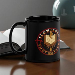 May include: Black ceramic mug with the words "SLOW BURN SMUT CLUB" in a circular design. The design includes an open book with flames and dripping liquid. The mug has a black handle and interior.