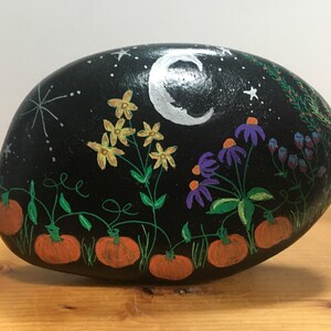 May include: A hand-painted, oval-shaped rock with a black background. The rock features a night scene with a crescent moon, stars, and various flowers. Orange pumpkins are painted at the bottom. The flowers are yellow, purple, and blue.