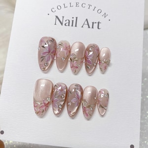 Op de afbeelding: Een set van tien roze en witte nageltips met bloemenmotieven en strass. De nageltips zijn in twee rijen op een witte kaart gerangschikt met de tekst "COLLECTION. Nail Art."