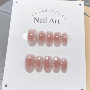 May include: Two sets of artificial nails are displayed on a white card. The nails are a light pink with a glittery silver base and white floral designs. The card reads "COLLECTION Nail Art."