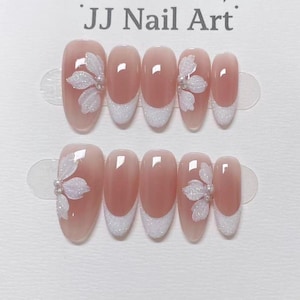 Könnte beinhalten: Zwei Sätze oval geformter Kunstnägel. Jeder Nagel ist hellrosa mit einer weißen Glitzer-French-Spitze. Weiße Glitzerblumen-Designs mit Perlenakzenten befinden sich auf einigen Nägeln. Der Text "JJ Nail Art" steht oben.