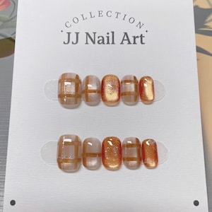 May include: A set of press-on nails with a grid pattern in shades of brown and gold. The nails are arranged on a white card with the text "COLLECTION JJ Nail Art". The nails have a glossy finish.