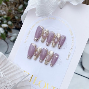 Peut inclure: Ensemble de dix faux ongles avec une base lavande, des accents argent&eacute;s et des embellissements en perles. Les ongles sont pr&eacute;sent&eacute;s sur une carte blanche avec le texte "I love you three thousand times" r&eacute;p&eacute;t&eacute; des deux c&ocirc;t&eacute;s.