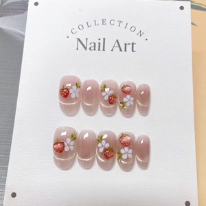 May include: A set of oval false nails with a light pink base and glitter. Each nail has a hand-painted design of a red strawberry and white flowers. The card reads "Collection Nail Art."