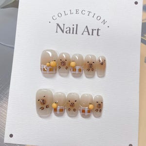 Può includere: Un cartoncino bianco con il testo "COLLECTION. Nail Art." stampato su di esso. Ci sono 7 piccole unghie finte quadrate bianche con un design di orso marrone e frutta arancione su uno sfondo a scacchi.