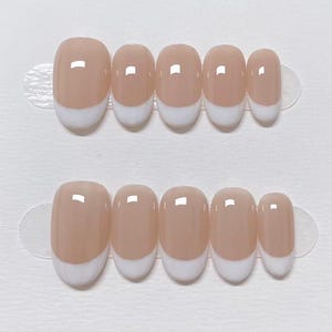 Pode incluir: Dois conjuntos de unhas postiças ovais. Cada unha é de cor bege claro com ponta francesa branca. As unhas estão dispostas em abas adesivas transparentes, prontas para aplicação. As unhas têm um acabamento brilhante.