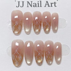 May include: Set of ten oval-shaped false nails. Each nail features a gradient design, transitioning from a nude pink to a gold glitter tip. A gold crisscross pattern is applied over the pink base. The text "JJ Nail Art" is at the top.