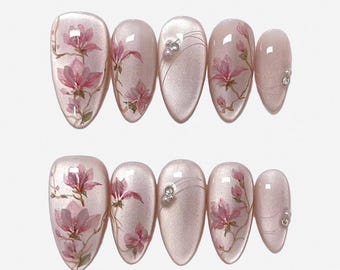 Könnte beinhalten: Ein Set aus zehn mandelförmigen Press-on-Nägeln mit einer schimmernden hellrosa Basis. Das Design zeigt handgemalte rosa Magnolienblüten auf braunen Zweigen, akzentuiert durch feine silberne Linien und kleine weiße Perlen auf ausgewählten Nägeln.