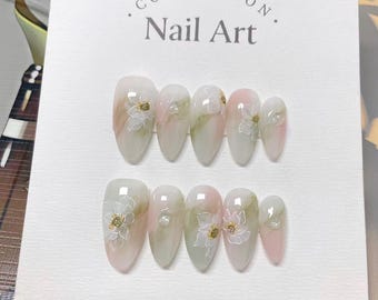 May include: A set of almond-shaped false nails with a floral design. The nails feature a gradient of pink, white, and green, with delicate white flowers and gold accents. The card behind the nails reads "COLLECTION. Nail Art."
