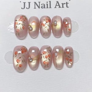 Puede incluir: Juego de uñas postizas ovaladas con base rosa claro, adornadas con diseños florales en naranja y verde. Cada uña presenta un adorno de perla. El texto "JJ Nail Art" está en la parte superior.
