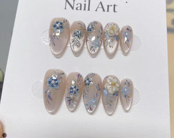 Puede incluir: Un conjunto de uñas postizas con un diseño floral. Las uñas son de color rosa claro con detalles de flores azules y blancas. El texto "Collection Nail Art" está impreso en la tarjeta.