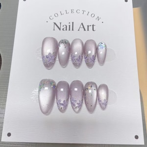 Pode incluir: Dois conjuntos de nail art cor lavanda, cada um com um design floral e detalhes iridescentes. As unhas são exibidas em um cartão branco com o texto "COLLECTION. Nail Art". As unhas têm formato oval.