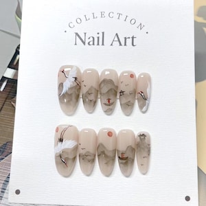 Op de afbeelding: Nail Art collectie met een set nepnagels. Elke nagel is beschilderd met een landschapsontwerp, inclusief kraanvogels, bergen en een ondergaande zon. De nagels zijn amandelvormig met een neutrale basiskleur.