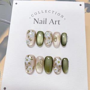 Pode incluir: Um conjunto de seis unhas postiças ovais com um design floral. As unhas são pintadas em tons de verde e bege com flores brancas e folhas verdes. O texto "COLLECTION. Nail Art." está impresso no cartão branco atrás das unhas.