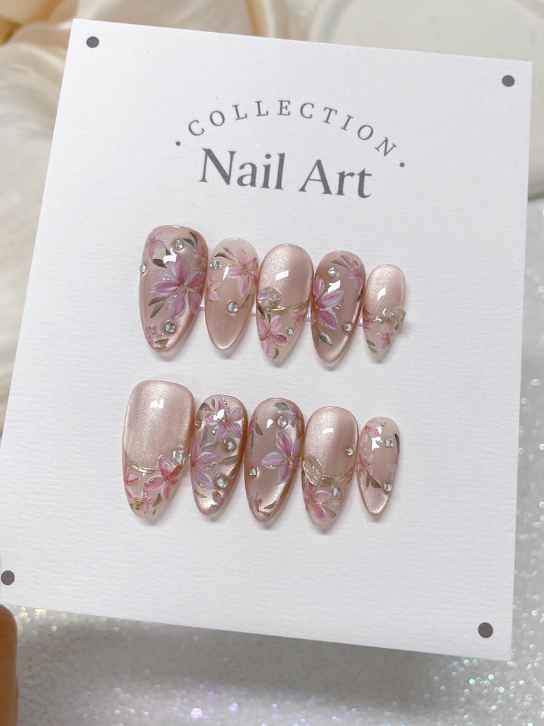 Op de afbeelding: Een set van tien roze en witte nageltips met bloemenmotieven en strass. De nageltips zijn in twee rijen op een witte kaart gerangschikt met de tekst "COLLECTION. Nail Art."