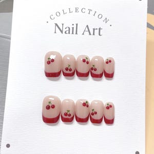 May include: A set of ten false nails with a cherry design. The nails are a light pink color with a red tip and a cherry design on each nail.