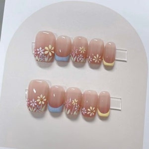 Puede incluir: Conjunto de diez uñas postizas con base rosa claro y diseño floral blanco y amarillo. Las uñas tienen un acento azul claro y amarillo en las puntas.