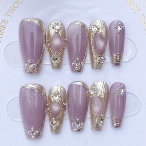 Peut inclure: Ensemble de dix faux ongles avec une base violet clair et des accents dor&eacute;s. Chaque ongle a un design diff&eacute;rent avec un petit strass transparent.
