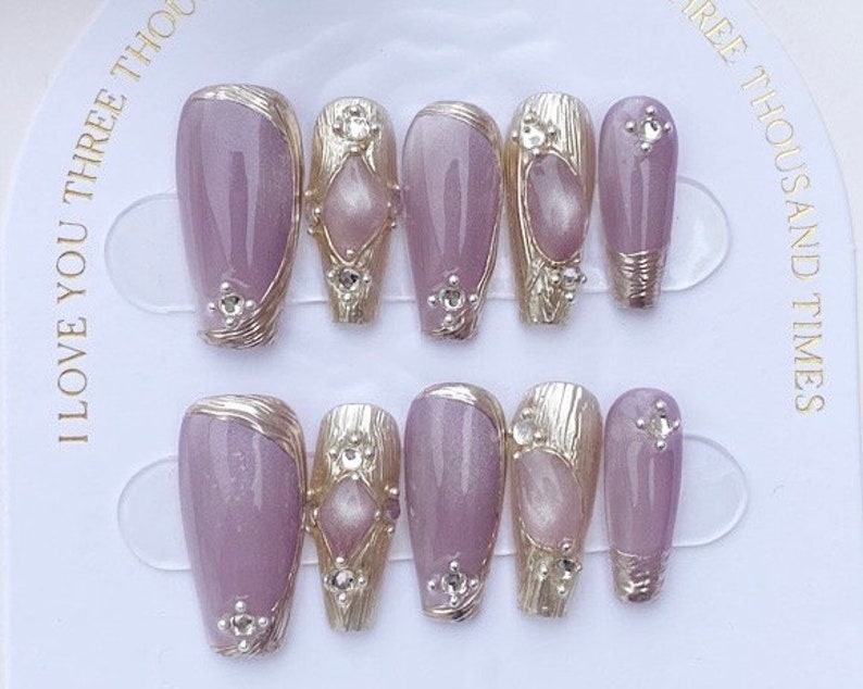 Peut inclure: Ensemble de dix faux ongles avec une base violet clair et des accents dor&eacute;s. Chaque ongle a un design diff&eacute;rent avec un petit strass transparent.