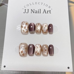 Peut inclure: Deux ensembles de faux ongles sont présentés sur une carte blanche. Les ongles présentent un motif avec des accents de bambou et de fleurs dans des tons de marron, blanc et or. Le texte "COLLECTION JJ Nail Art" est imprimé en haut.