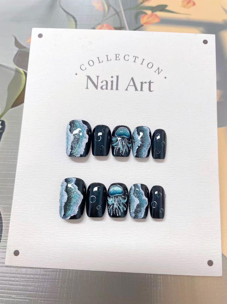 Peut inclure: Ensemble de faux ongles avec une base bleu fonc&eacute;. Certains ongles pr&eacute;sentent un motif marbr&eacute; blanc et bleu sarcelle, tandis que d'autres pr&eacute;sentent une illustration de m&eacute;duse. Le texte "Collection Nail Art" est visible.