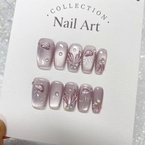 May include: A set of ten light pink artificial nails with a jellyfish design. The nails are decorated with iridescent glitter and small white dots. The nails are arranged in two rows on a white card with the text "COLLECTION Nail Art."