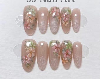May include: A set of ten artificial nails with a floral design. The oval-shaped nails are painted in a nude colour. Each nail features hand-painted flowers in shades of pink, peach, and green, with gold accents. The text "JJ Nail Art" is at the top.