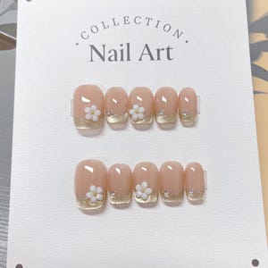 May include: A set of ten nude-colored press-on nails with gold accents and white flower embellishments. The nails are displayed on a white card with the text "COLLECTION. Nail Art."