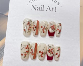 May include: A set of ten false nails with a white base and a design of red berries and green leaves. The nails are arranged in two rows of five. The text "COLLECTION. Nail Art." is printed on the white card the nails are displayed on.