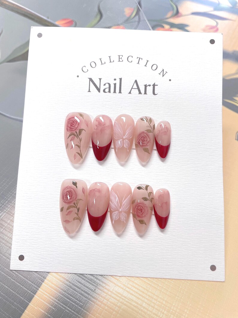 May include: A set of ten fake nails with a floral design. The nails are a light pink color with a red tip. The floral design features pink roses and butterflies.