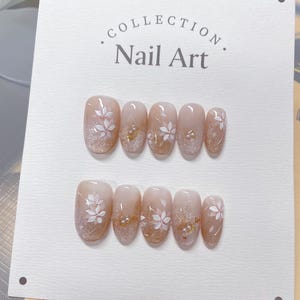May include: A set of ten oval-shaped false nails with a light brown base and white floral designs. The nails have a glittery finish and some have small gold beads.