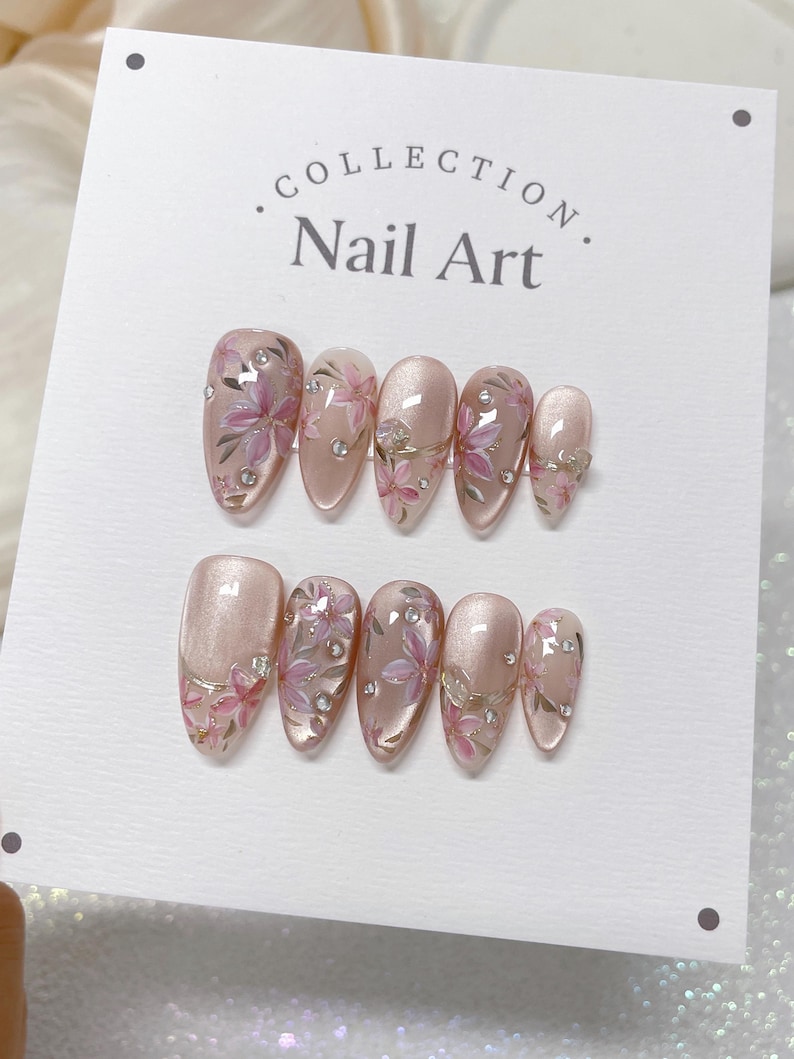 Op de afbeelding: Een set van tien roze en witte nageltips met bloemenmotieven en strass. De nageltips worden getoond op een witte kaart met de tekst "COLLECTION. Nail Art."