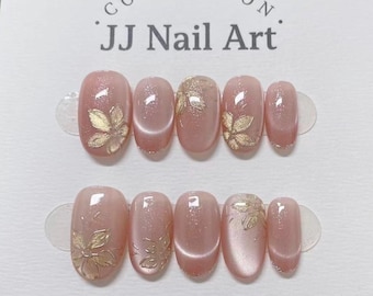 Könnte beinhalten: Ein Set aus zehn künstlichen Fingernägeln mit schimmerndem rosa Katzenaugen-Effekt und goldenen Blumenakzenten. Die Nägel sind in zwei Reihen auf einer weißen Karte mit der Aufschrift COLLECTION JJ Nail Art angeordnet und haben eine abgerundete Form.