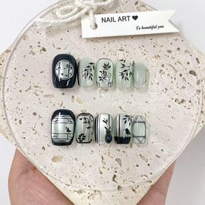 May include: A set of press-on nails with various designs. Some nails are black with white accents, while others are light green with black floral patterns. The tag reads "NAIL ART" and "To beautiful you".