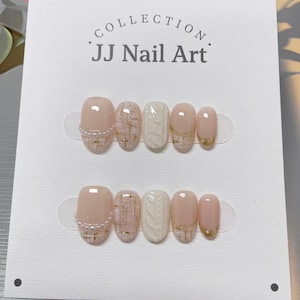 Puede incluir: Dos juegos de uñas artificiales se muestran en una tarjeta blanca. Las uñas presentan una variedad de diseños, que incluyen una base rosa claro con detalles dorados, una uña blanca texturizada y un borde perlado. La tarjeta dice "COLLECTION JJ Nail Art."