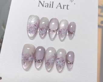 May include: A set of almond-shaped false nails with a floral design. The nails are a mix of clear, white, and lavender, with delicate purple flower accents and small rhinestones. The card reads "COLLECTION Nail Art."