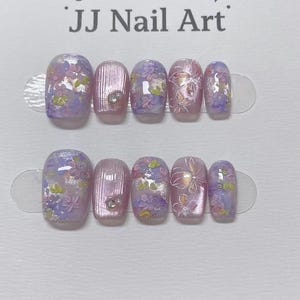 Könnte beinhalten: Zwei Sets von Press-On-Nägeln mit floralem Design. Die Nägel haben eine hellviolette Basis mit rosa, lila und grünen floralen Akzenten. Einige Nägel haben einen kleinen Edelstein. Der Text "JJ Nail Art" steht oben.