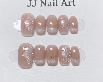 Könnte beinhalten: Zwei Sets künstlicher Nägel mit hellrosa Basis. Jeder Nagel hat ein weißes Blumendesign, einen Perlenakzent und goldene Glitzerdetails. Der Text "JJ Nail Art" steht oben.