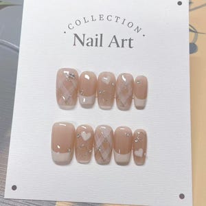 Può includere: Un set di unghie finte con una palette di colori neutri. Le unghie presentano un design a punta francese, accenti a cuore e a quadri e dettagli glitterati argentati. La carta dietro le unghie recita "Collection Nail Art."