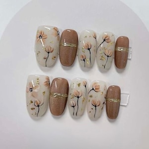 May include: Set of ten oval-shaped press-on nails with a floral design. The nails are painted with a white base and feature delicate brown flowers with gold accents. Some nails have a solid brown color with a gold accent.
