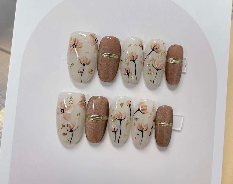 May include: Set of ten oval-shaped press-on nails with a floral design. The nails are painted with a white base and feature delicate brown flowers with gold accents. Some nails have a solid brown color with a gold accent.