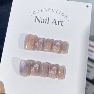 May include: A set of ten press-on nails with a light pink base and iridescent glitter. Each nail has a white cloud design and a small silver accent.