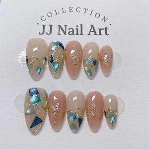 May include: A set of ten press-on nails from the JJ Nail Art collection, featuring a nude base with geometric blue and silver foil accents, gold bead embellishments, and fine gold line detailing arranged in two rows on a white display card.