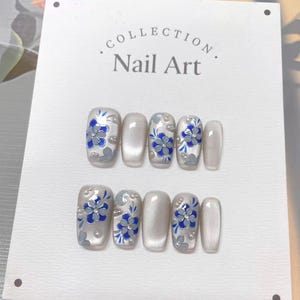 May include: A set of false nails with a floral design. The nails are pearlescent white with blue and grey flower accents and small pearl embellishments. The text "Collection Nail Art" is at the top.