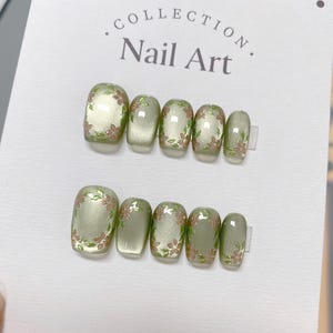Puede incluir: Conjunto de diez puntas de uñas verdes y marrones con diseños florales. Las puntas de uñas están dispuestas en dos filas de cinco. El texto "COLLECTION. Nail Art." está impreso en el fondo blanco.