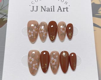 May include: Two sets of almond-shaped false nails. Each nail is painted in shades of brown and beige, with floral designs and gold leaf accents. The card reads "COLLECTION" and "JJ Nail Art".