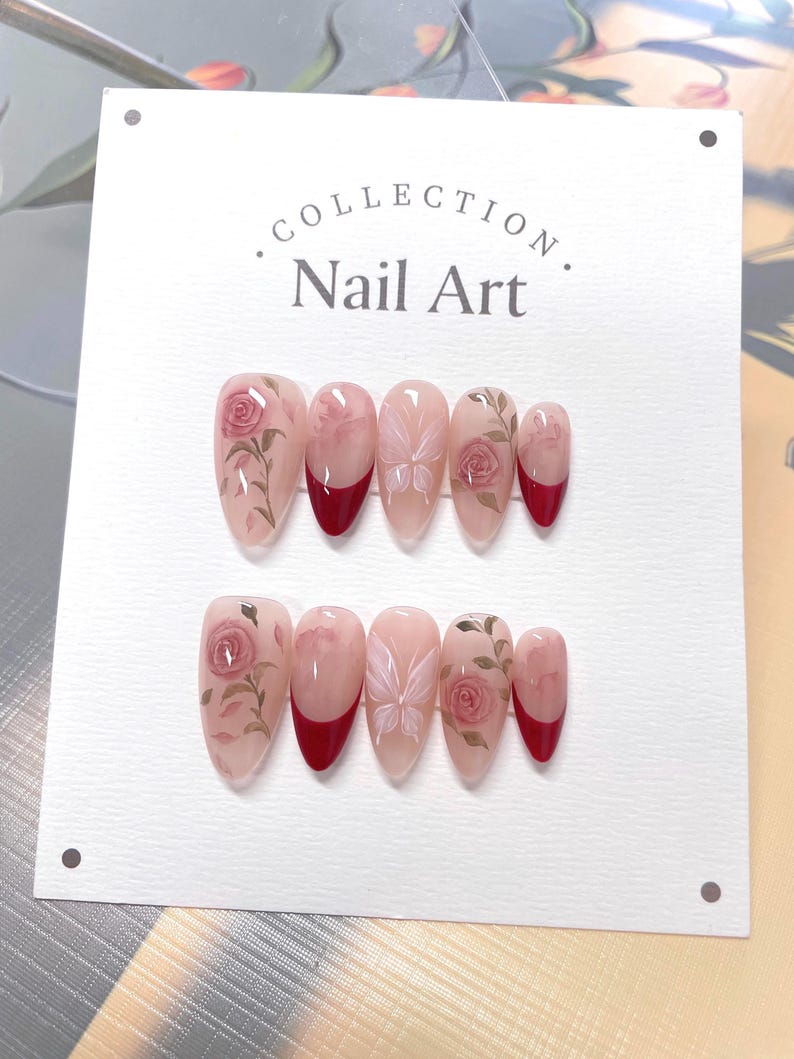 May include: A set of ten fake nails with a floral design. The nails are a light pink color with a red tip. The floral design features pink roses and butterflies.