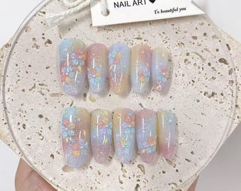 Op de afbeelding: Een set van tien kunstnagels met een bloemenontwerp. De nagels hebben een verloop van pastelkleuren, waaronder roze, geel en blauw, met glitteraccenten. De tekst "NAIL ART" staat op een label.